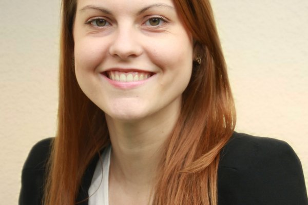 Foto von Elisabeth Huther vor einem neutralen Hintergrund. Sie hat lange, rotbraune Haare und trägt einen schwarzen Blazer. Sie lächelt in die Kamera.
